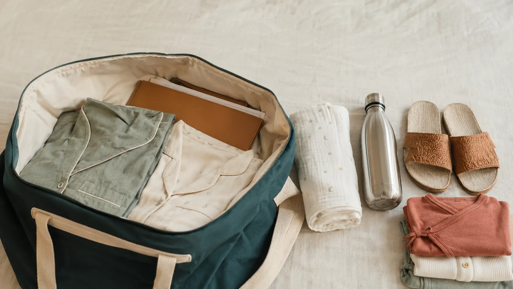 An open hospital bag packed for delivery, with folded cotton nightwear, a document folder, a swaddle blanket and a water bottle laid out beside it